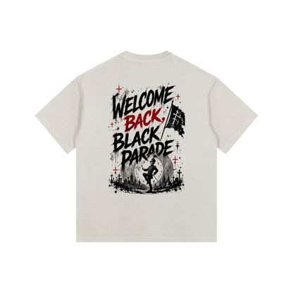 A light beige crewneck t-shirt shown from the back, featuring a bold graphic print with the text 'WELCOME BACK, BLACK PARADE' in black and red, accented with red crosses and a black-and-white flag illustration.