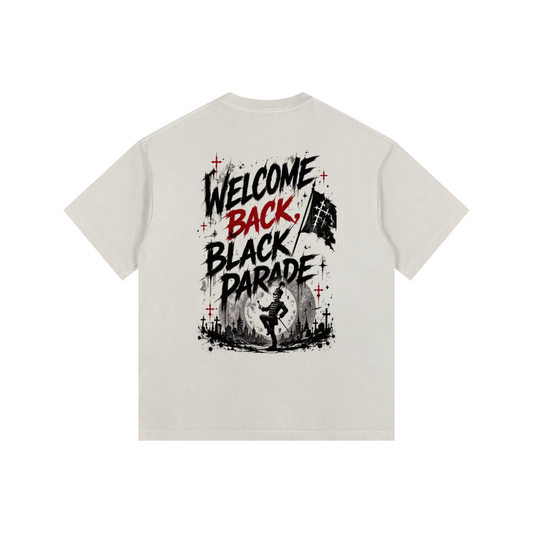 A light beige crewneck t-shirt shown from the back, featuring a bold graphic print with the text 'WELCOME BACK, BLACK PARADE' in black and red, accented with red crosses and a black-and-white flag illustration.