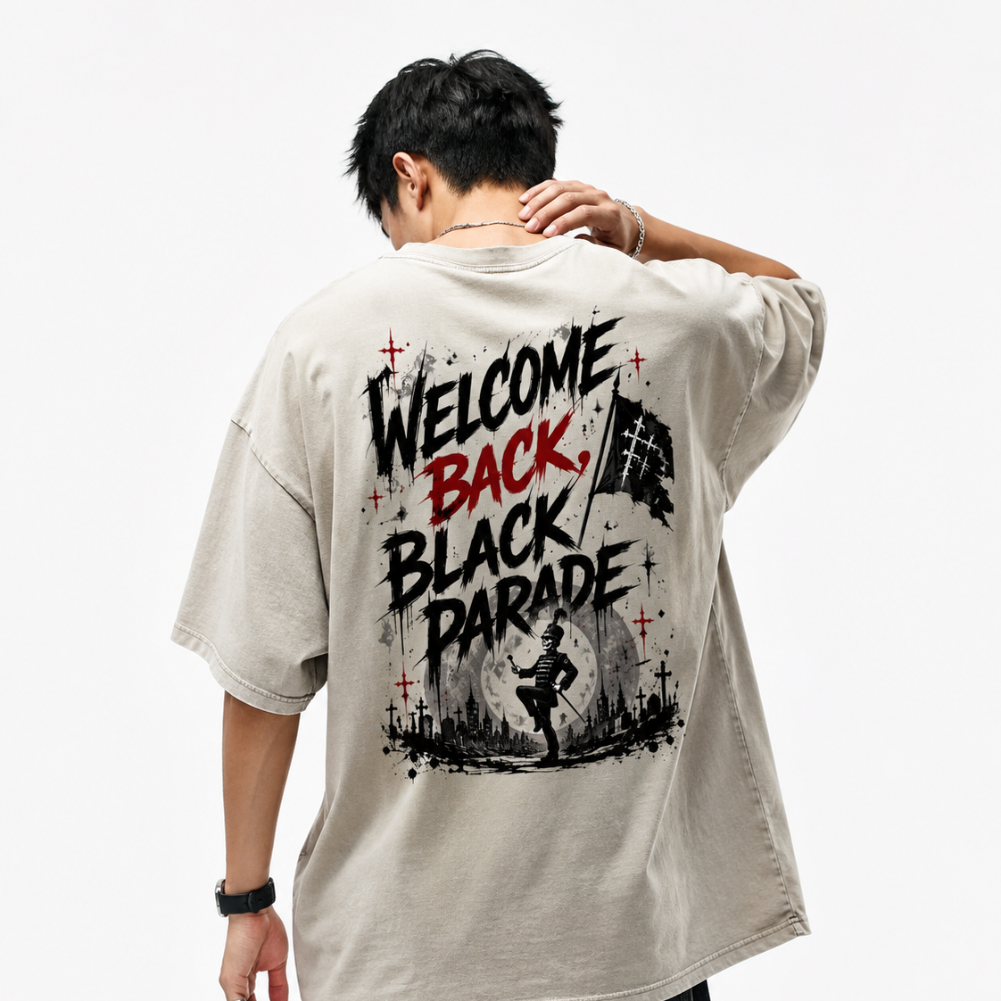 A man viewed from behind wearing an oversized light beige graphic T-shirt with a large back print reading 'WELCOME BACK, BLACK PARADE' in black and red, paired with dark jeans against a plain white studio background.
